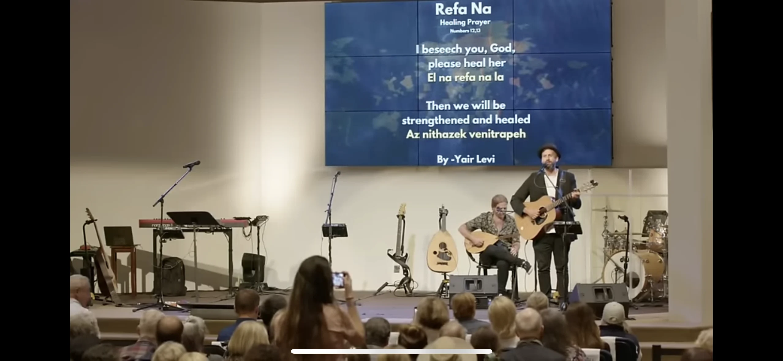 Yair singing Refa Na with a guitarist in front of a seated community gathering